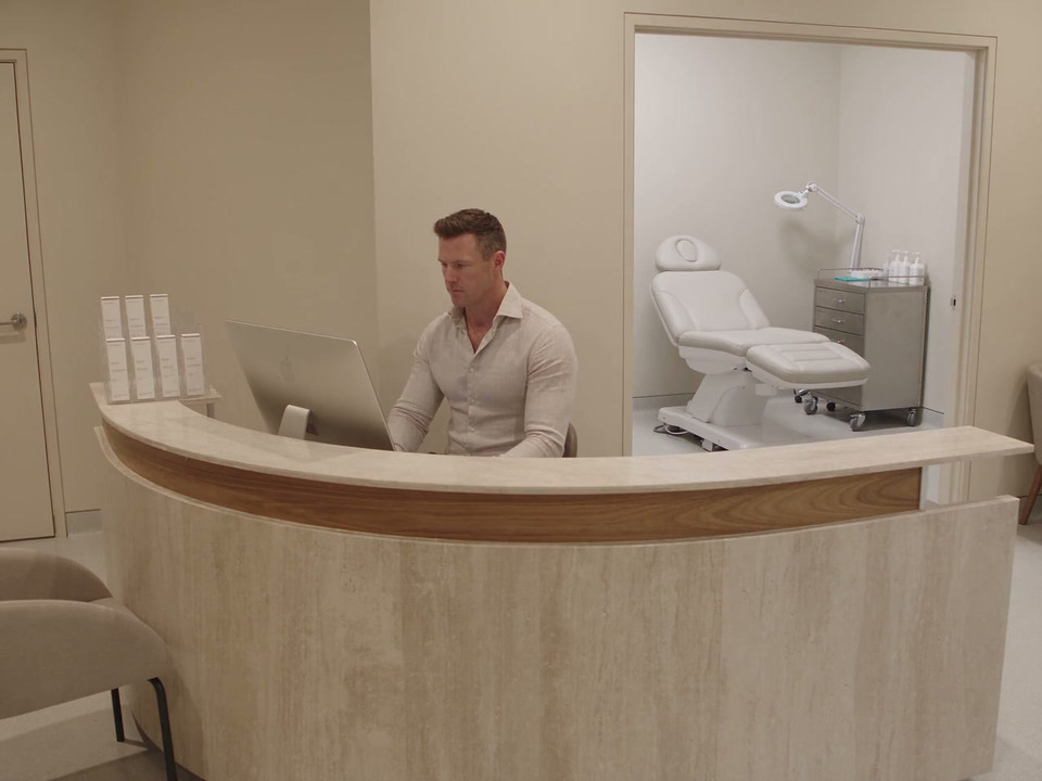 A clinic coordinator works at the reception computer, with a treatment room visible behind him